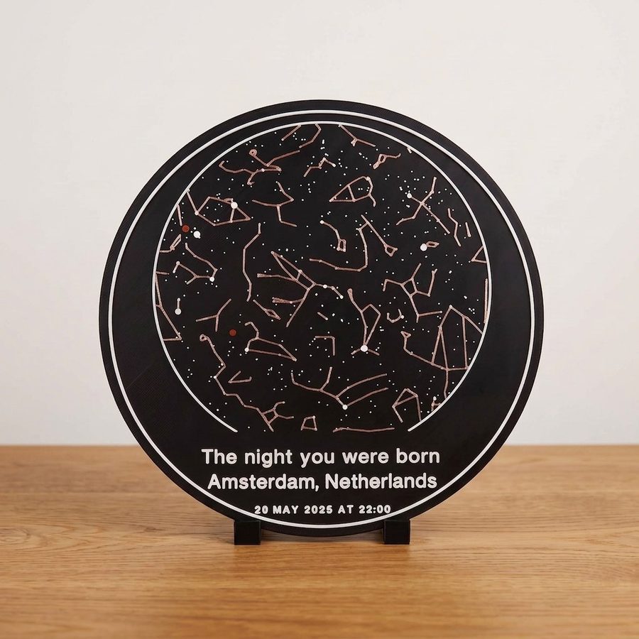 A round OrbitStudio Sky star map plate on a small black desk stand: copper constellation lines, white stars and two red planet dots on a dark plate, lettered The night you were born, Amsterdam, Netherlands, 20 May 2025 at 22:00